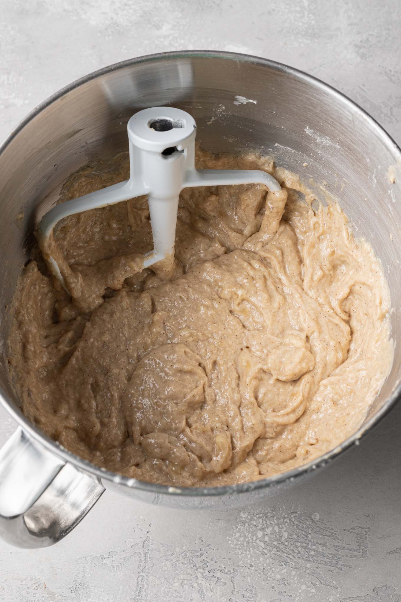 An overhead view of banana bread batter in a mixing bowl, with a paddle attachment from a stand mixer. 