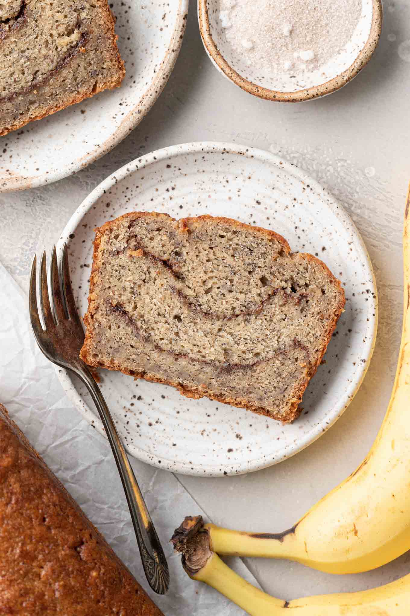 An overhead view of a slice of banana cinnamon bread on a dessert plate, with a fork. 