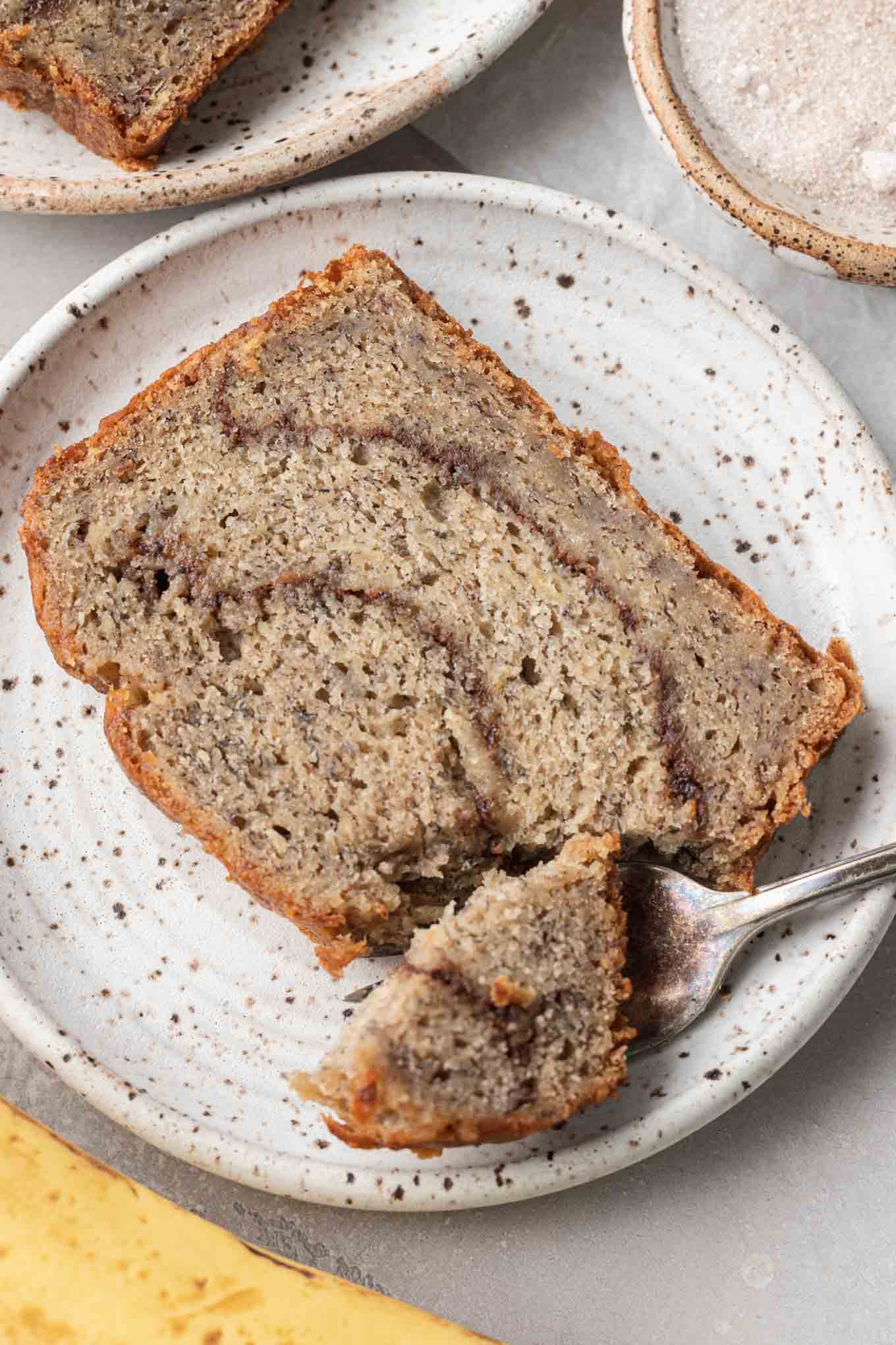 A close up view of a slice of cinnamon banana bread on a dessert plate. A piece of bread is speared on a fork. 