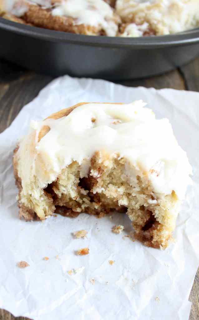 A 1 hour cinnamon roll with a bite missing on a piece of paper. A pan of cinnamon rolls rests in the background. 