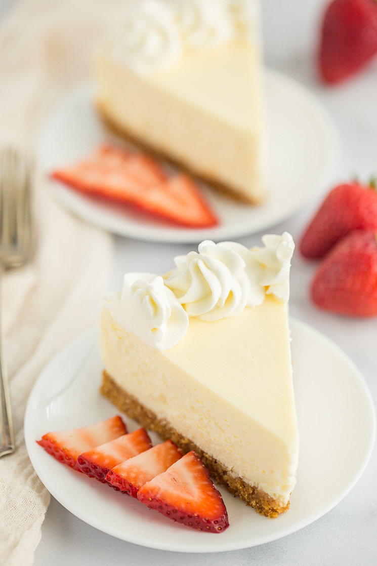 Two slices of cheesecake on white plates with sliced strawberries on the side of each one.