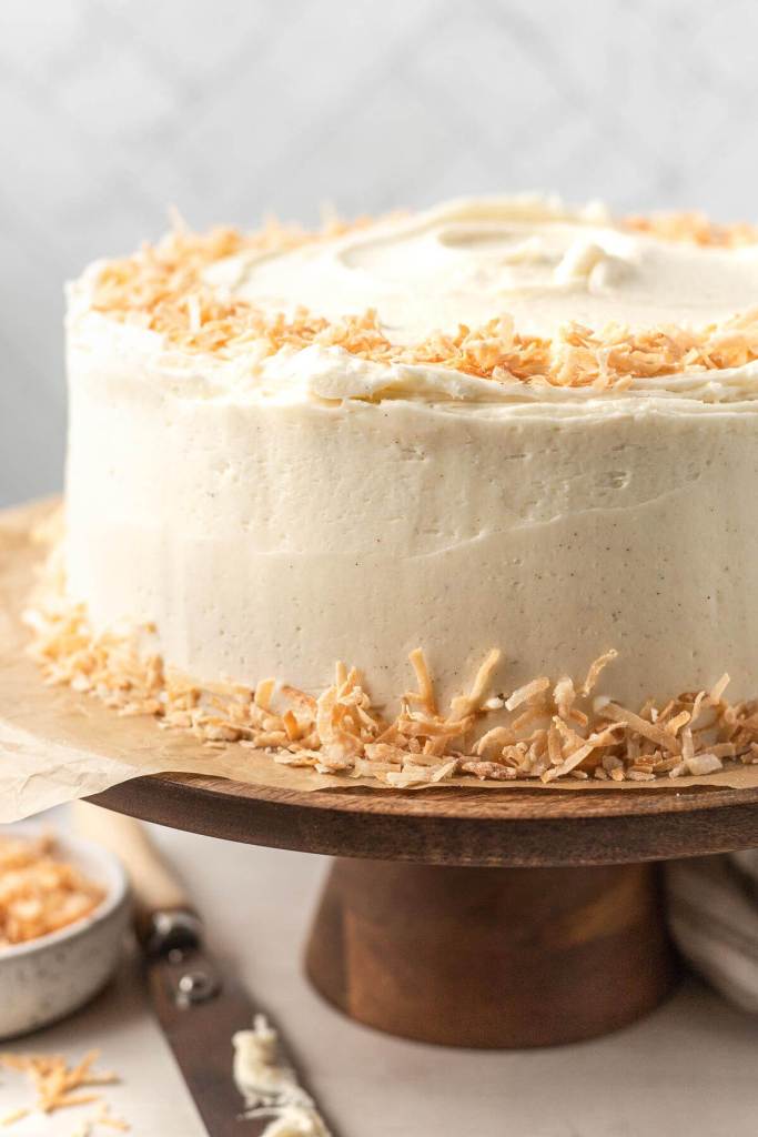 A coconut cake with coconut buttercream frosting and toasted coconut on a wooden cake stand. 