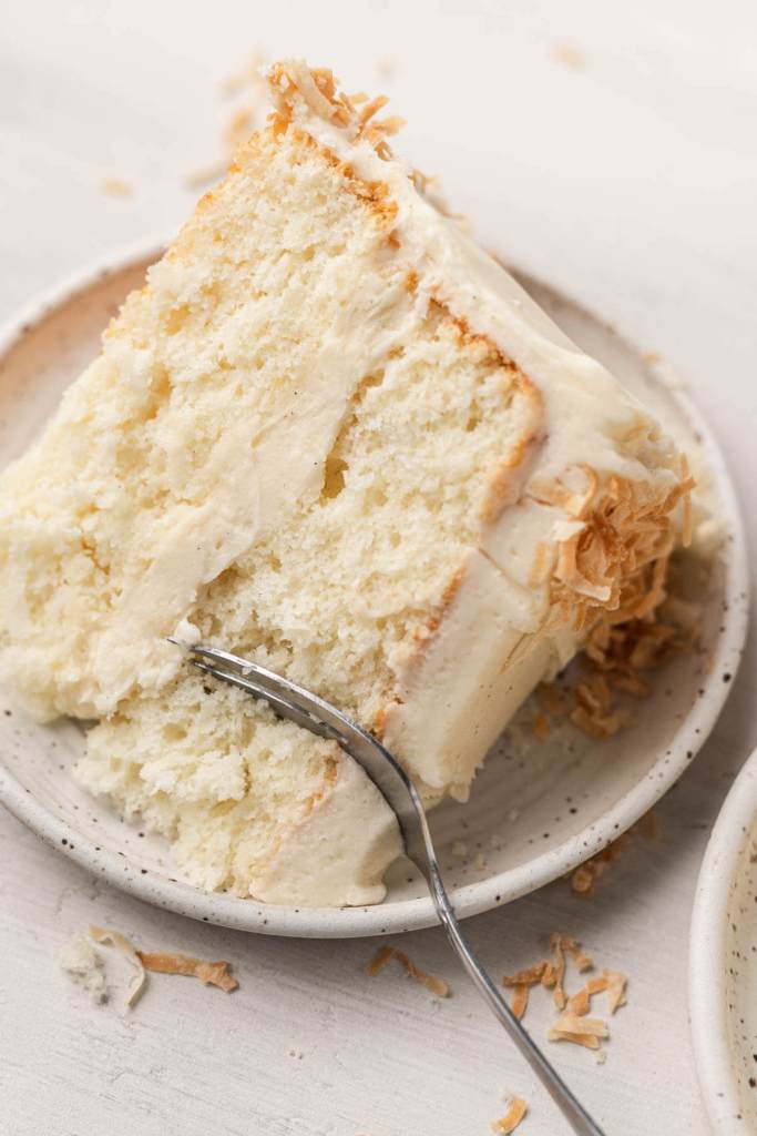 A piece of frosted coconut layer cake on a white speckled dessert plate with a fork. 