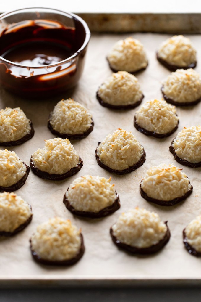 Chocolate dipped macaroons on a baking sheet. A dish of melted chocolate is also on the sheet.