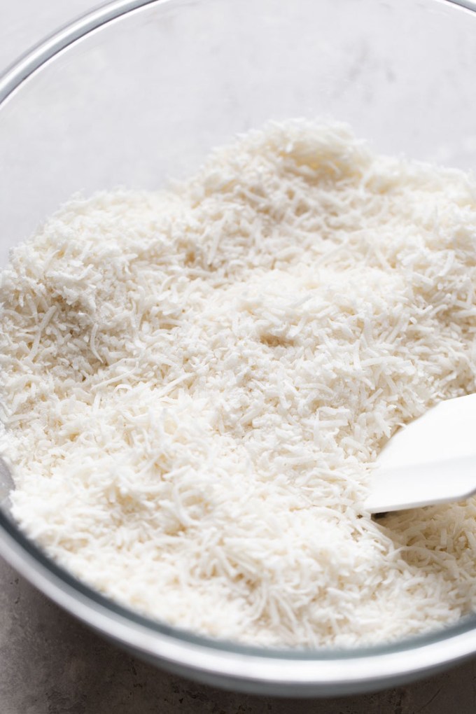 Sweetened shredded coconut in a glass mixing bowl with a rubber spatula.