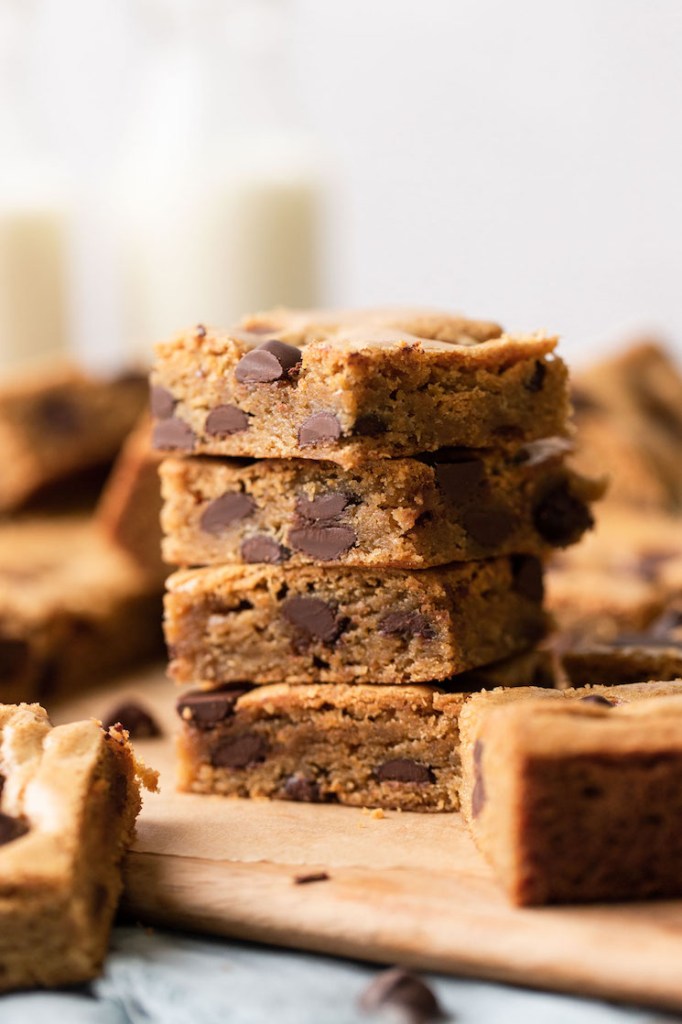 A stack of four chocolate chip cookie bars on a wood serving board. More cookie bars surround the stack. 