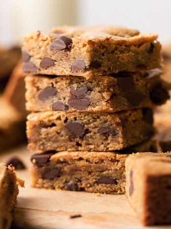 A stack of four chocolate chip cookie bars on a wood serving board. More cookie bars surround the stack.