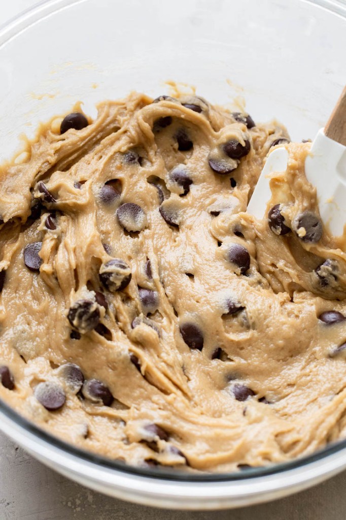 Raw chocolate chip cookie dough in a glass bowl with a rubber spatula. 