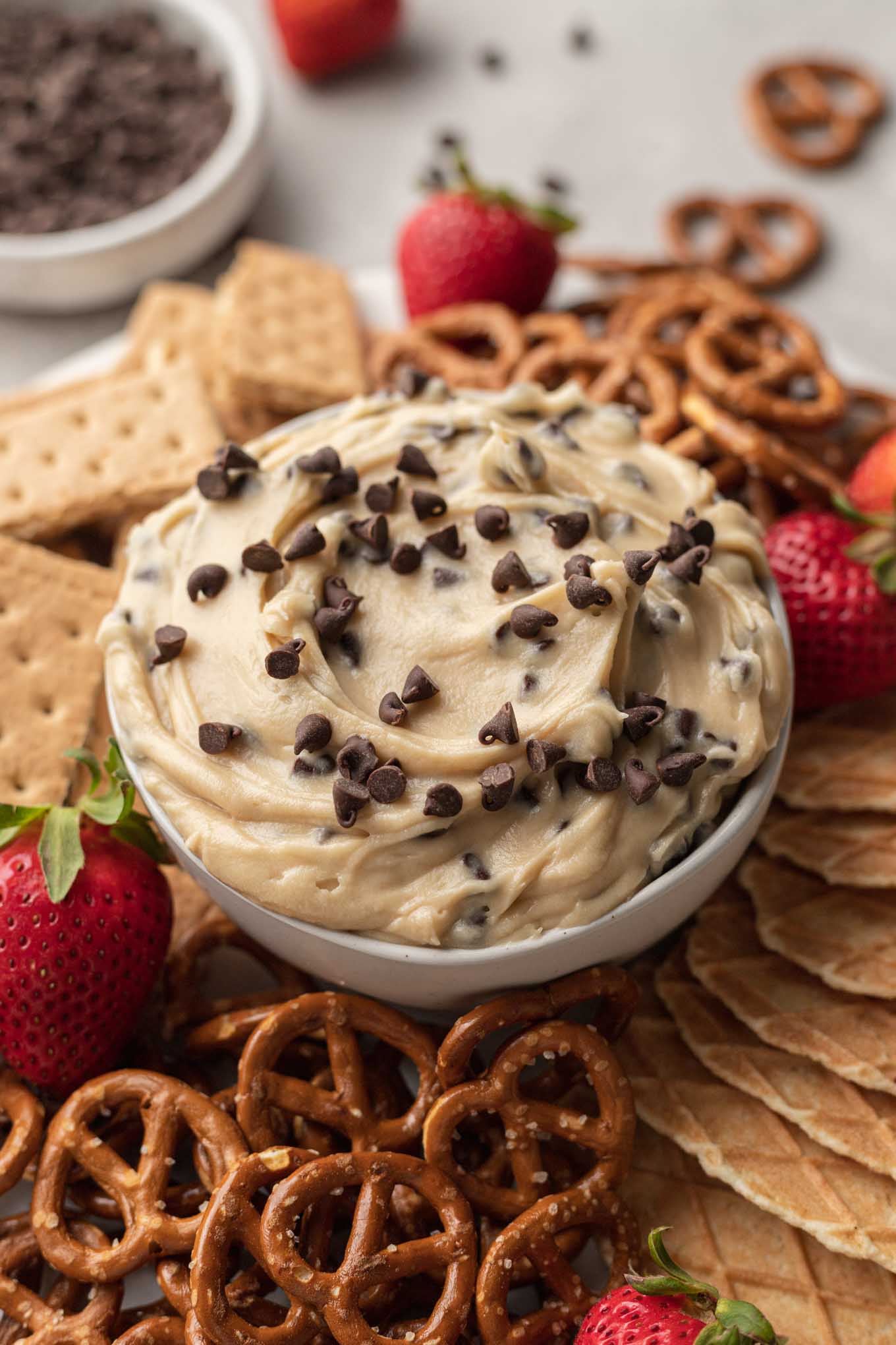 A dish of cream cheese cookie dough dip, surrounded by waffle cone pieces, pretzels, berries, and graham crackers. 