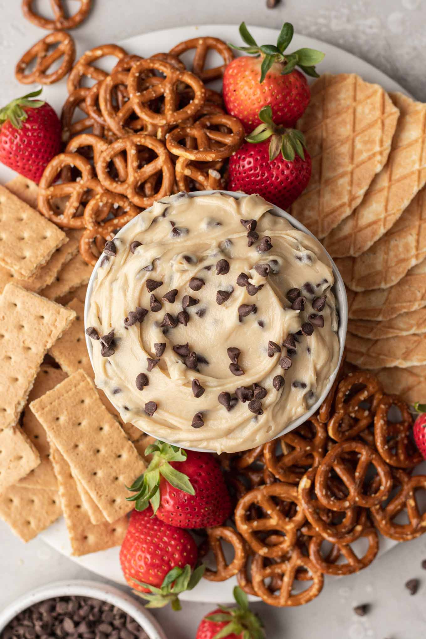 An overhead view of a dish of chocolate chip cookie dough dip on a platter with pretzels, graham crackers, and berries. 
