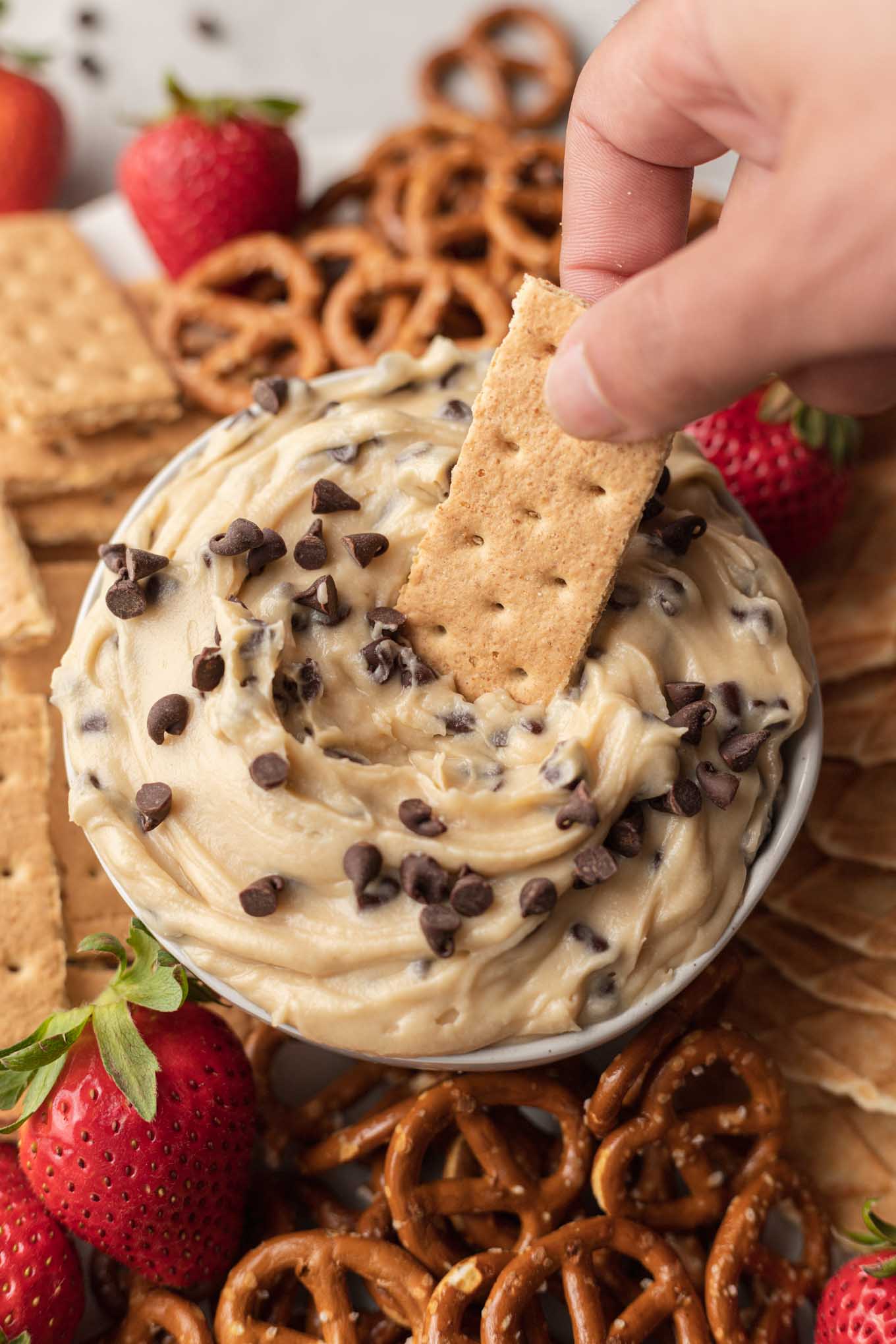 A graham cracker being dipped into chocolate chip cookie dough dip, which rests on a platter. 