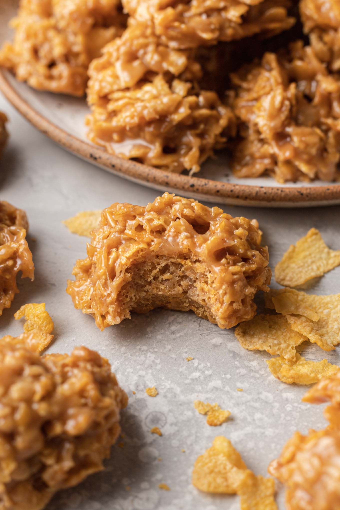 A close-up view of a cornflake cookie, with a bite missing. Additional cookies surround the central cookie. 