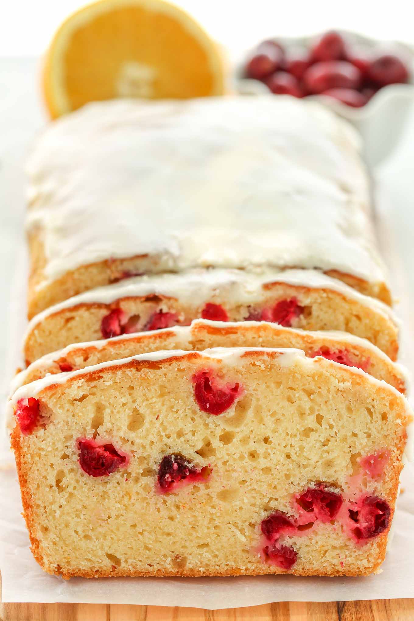 A loaf of cranberry orange bread topped with glaze. Half an orange and a bowl of cranberries rest in the background. 