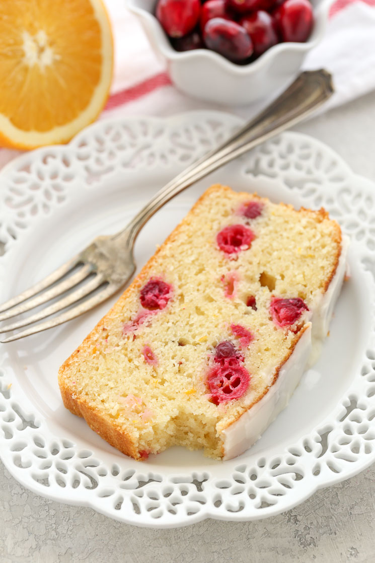 A slice of glazed cranberry orange bread on a white plate with a fork. Half an orange and a bowl of cranberries rest in the background. 