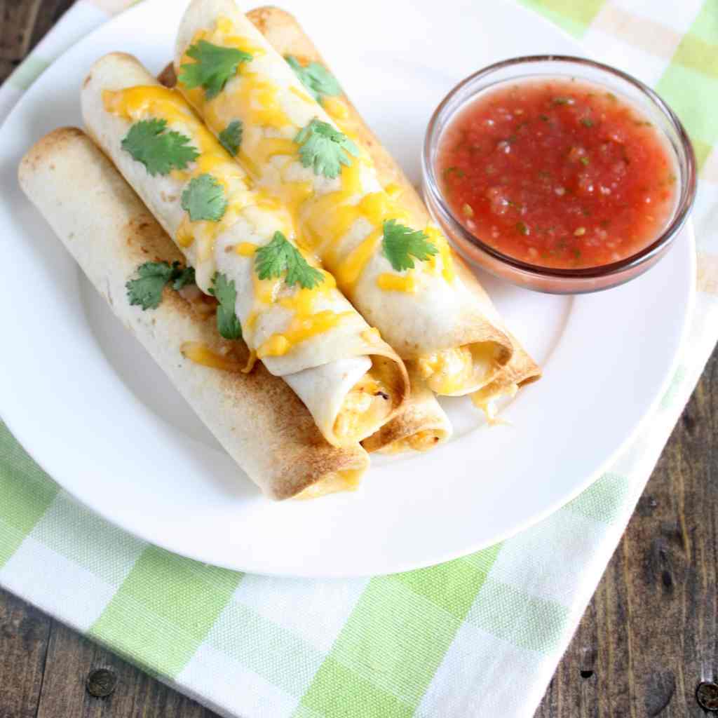 Overhead view of five baked chicken and cheese taquitos on a white plate with a dish of salsa. 