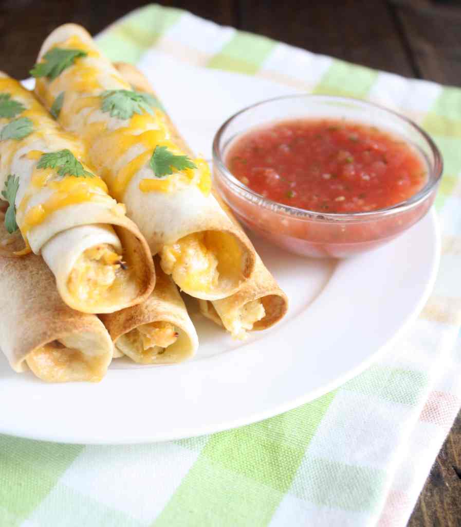 Five baked chicken taquitos on a white plate with a dish of salsa. 