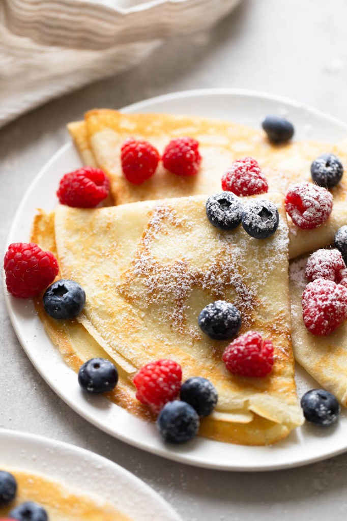 Three crepes topped with berries and dusted with powdered sugar on a white plate. A towel is in the background. 