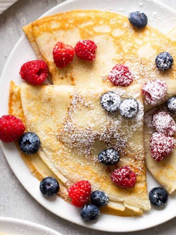 Overhead view of three homemade crepes topped with fresh berries and dusted with powdered sugar on a white plate.