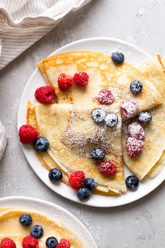 Overhead view of three homemade crepes topped with fresh berries and dusted with powdered sugar on a white plate. Another plate of crepes is partially seen in the lower left corner. 