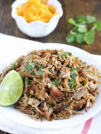 Shredded carnitas meat sitting on a white plate with cilantro and cheese in the background.