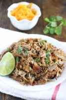 Shredded carnitas meat sitting on a white plate with cilantro and cheese in the background.