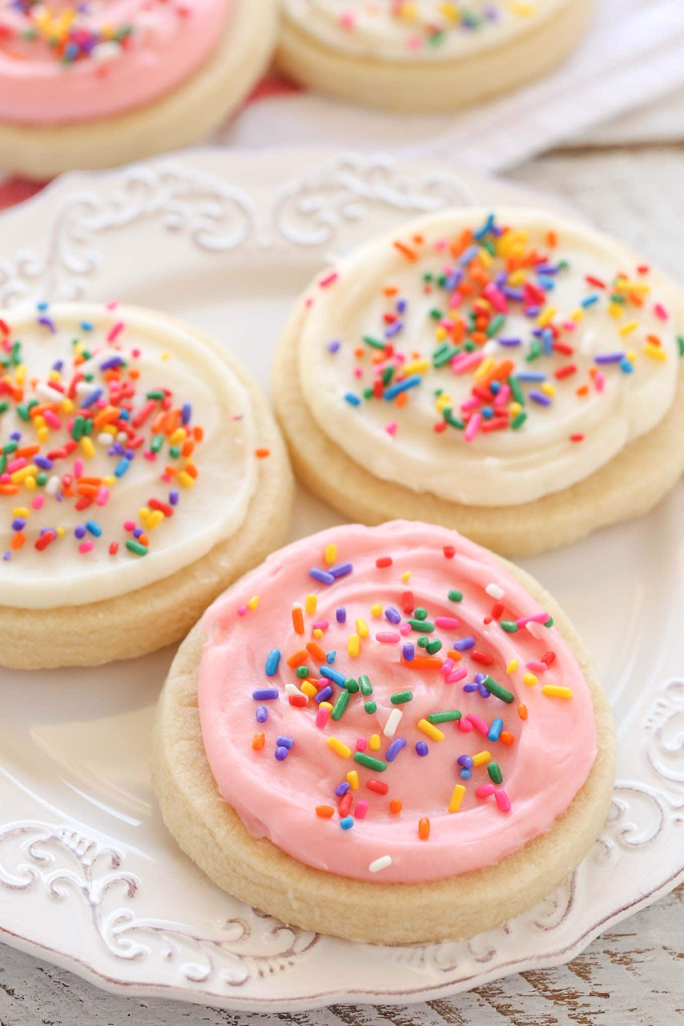 Three frosted sugar cookies on a white plate. Additional sugar cookies rest in the background. 