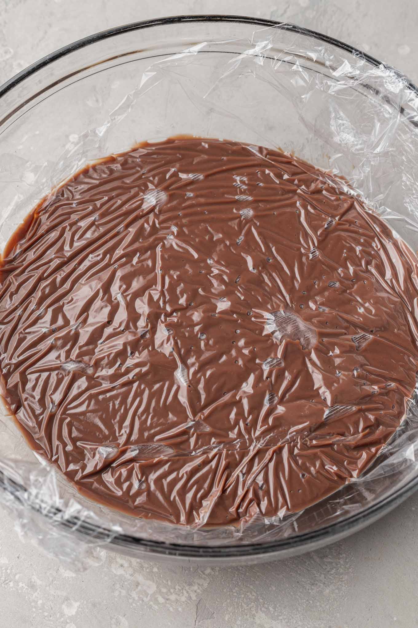 An overhead view of homemade chocolate pudding in a glass mixing bowl, with plastic wrap pressed on top. 