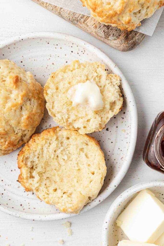 An overhead view of homemade drop biscuits on a white plate. One biscuit has been halved and topped with butter. 