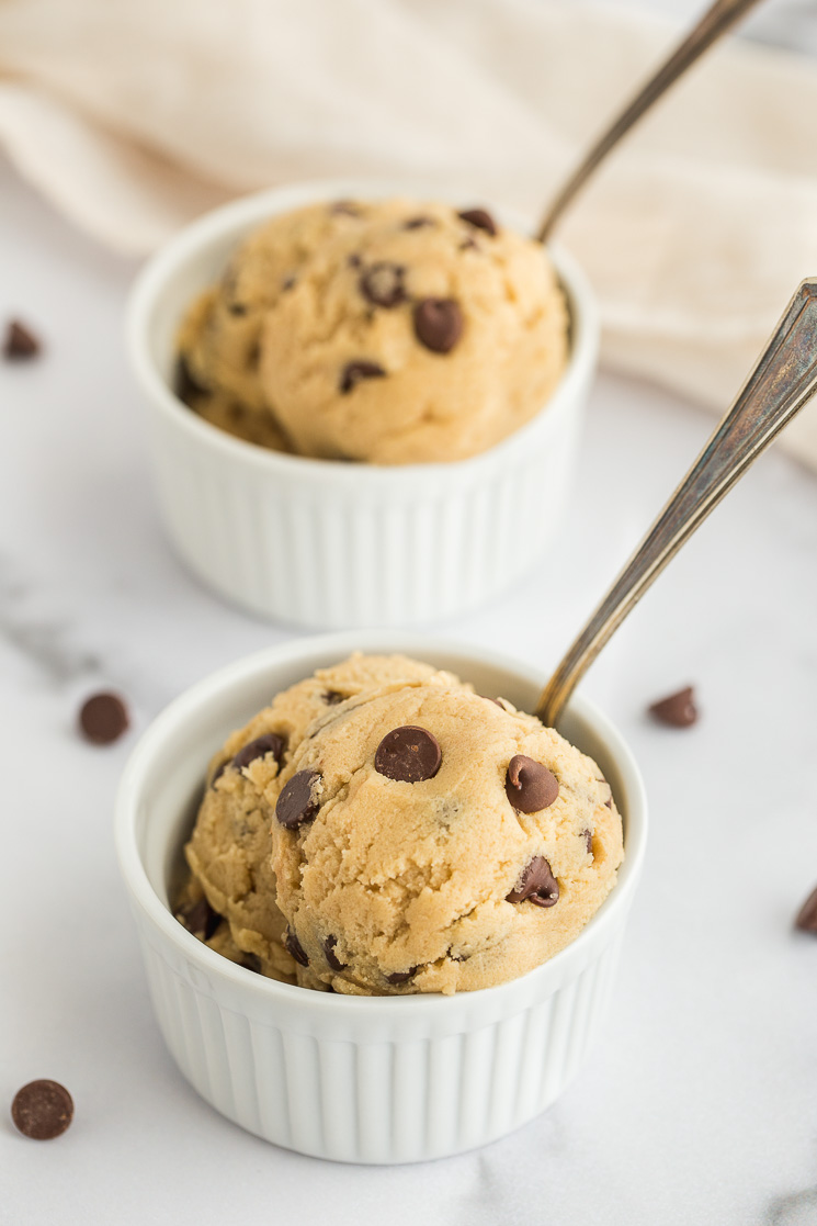 Two small white ramekins filled with cookie dough and a spoon in each one.