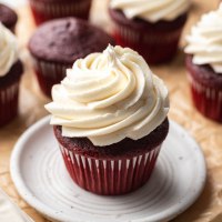 A red velvet cupcake topped with ermine frosting on a white plate. More cupcakes are around the plate.