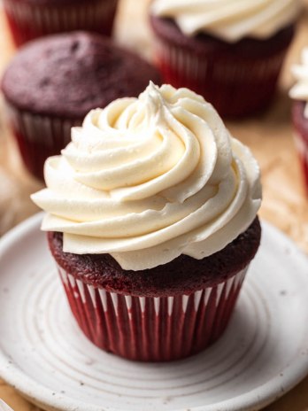 A red velvet cupcake topped with ermine frosting on a white plate. More cupcakes are around the plate.