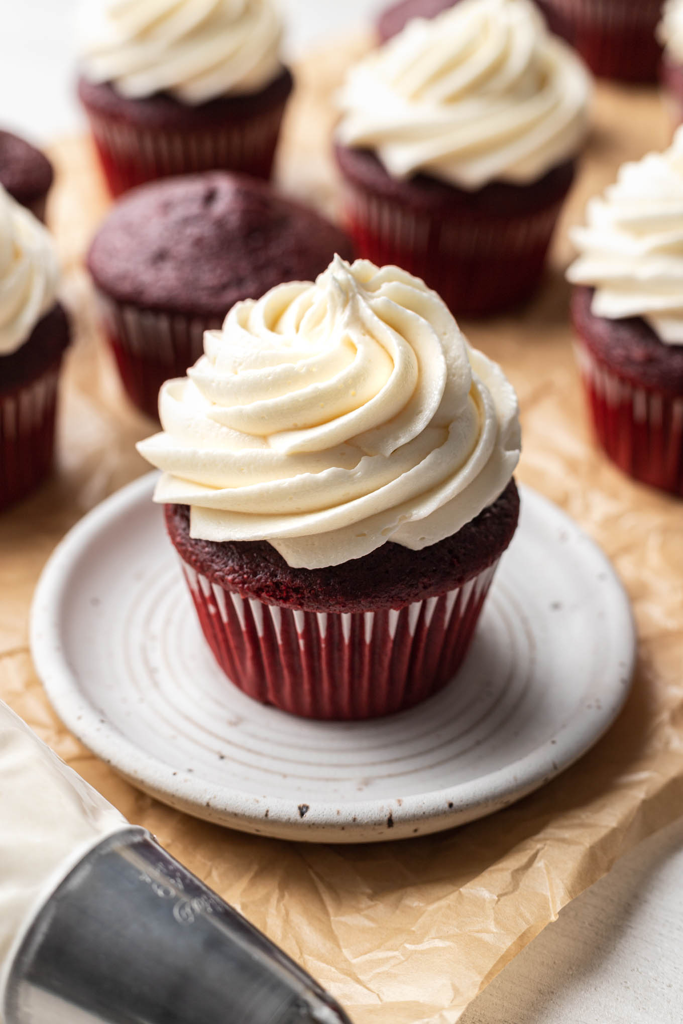 A red velvet cupcake topped with ermine frosting on a white dessert plate. More cupcakes surround the plate. 