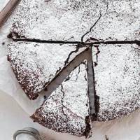 An overhead view of a flourless chocolate cake topped with powdered sugar. Two slices have been cut from the cake and one is being removed.