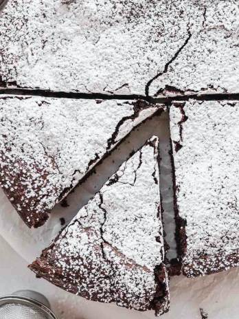 An overhead view of a flourless chocolate cake topped with powdered sugar. Two slices have been cut from the cake and one is being removed.