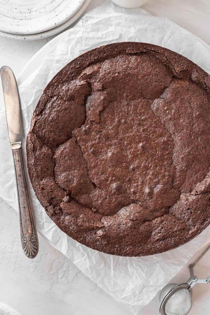 An overhead view of a flourless chocolate cake on a square of parchment paper. A butter knife rests on the side. 