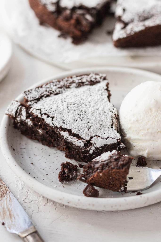 A slice of flourless cake dusted with powdered sugar on a white plate with a scoop of ice cream. 