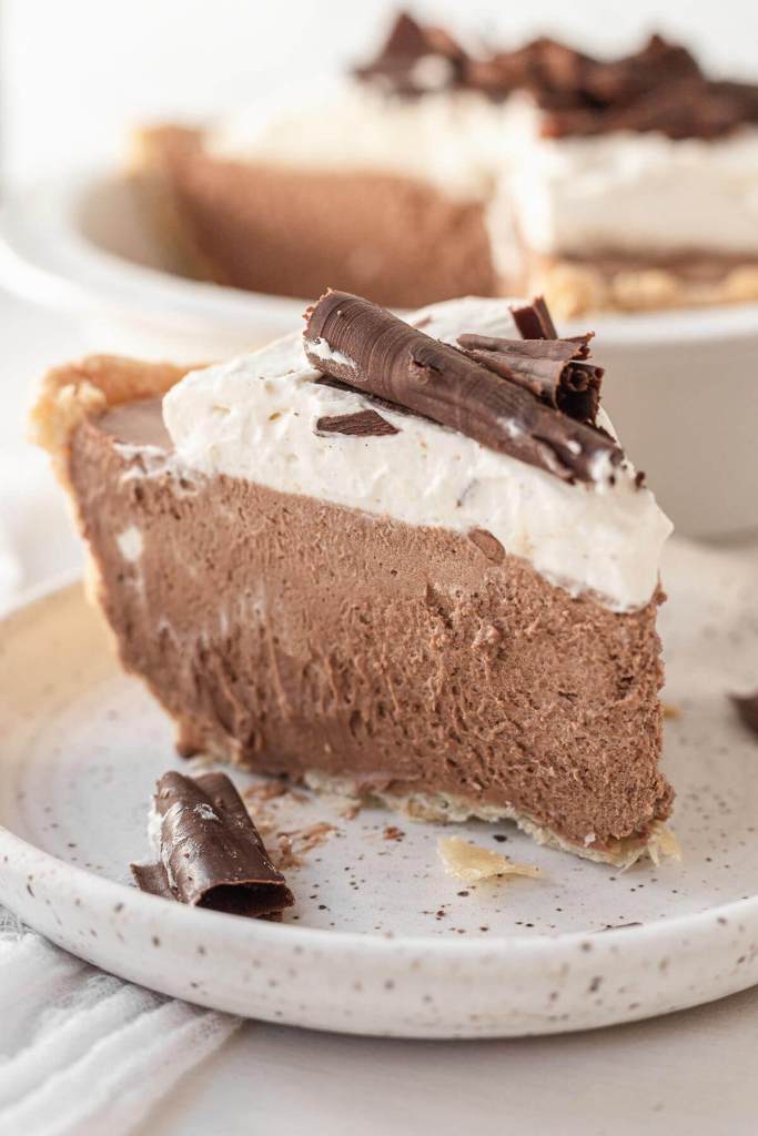 A side view of a slice of French silk chocolate pie on a speckled white plate. The rest of the pie is in a pie plate in the background. 