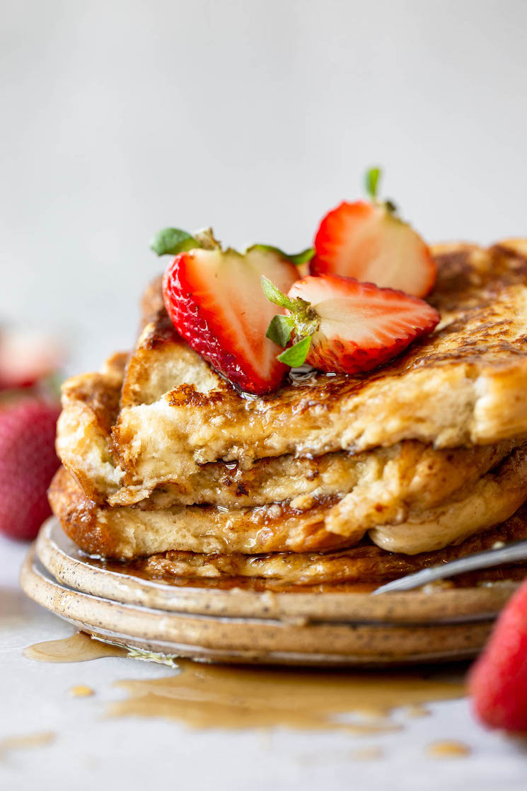 A plate holding French toast stacked up and topped with syrup and halved strawberries.