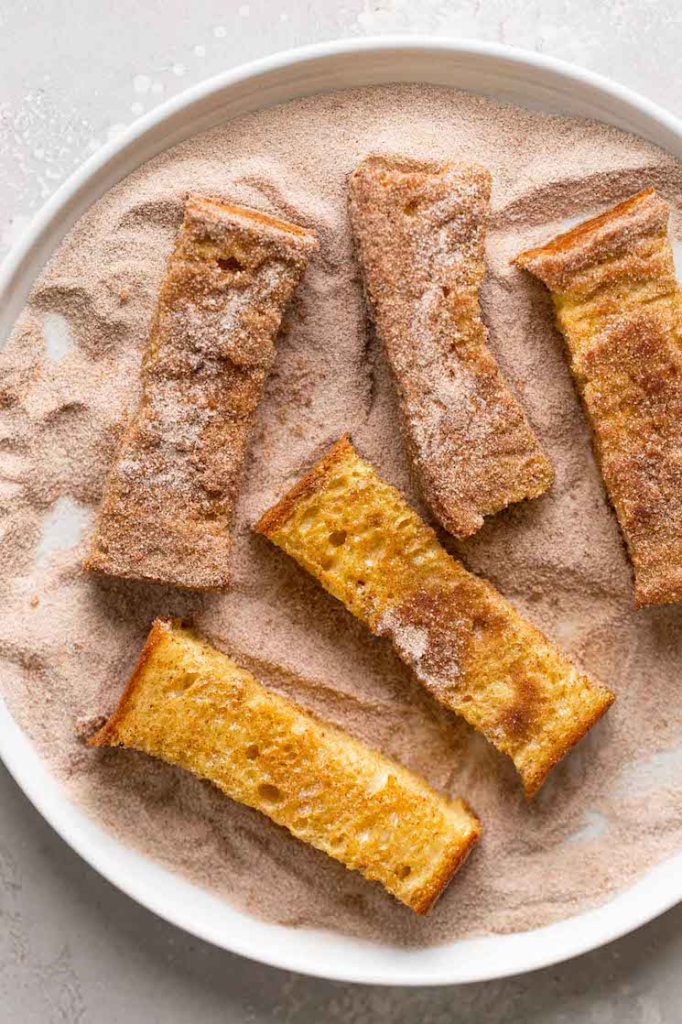 Cinnamon french toast sticks being rolled in cinnamon sugar, viewed from above. 