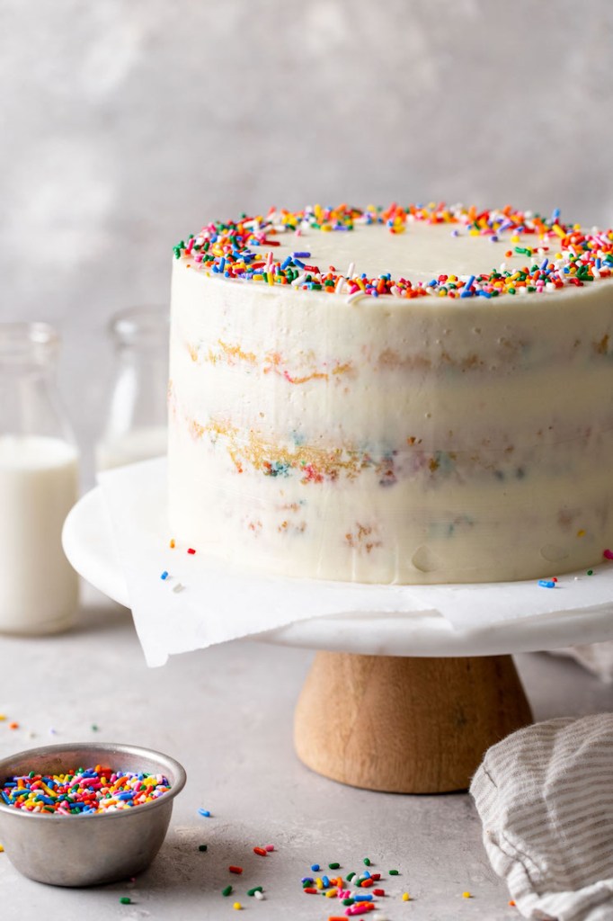 A homemade funfetti cake on a cake stand. Two glass bottles of milk are in the background and a dish of sprinkles is in the foreground. 