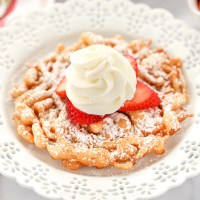 A funnel cake topped with strawberries and homemade whipped cream sitting on a decorative white plate.