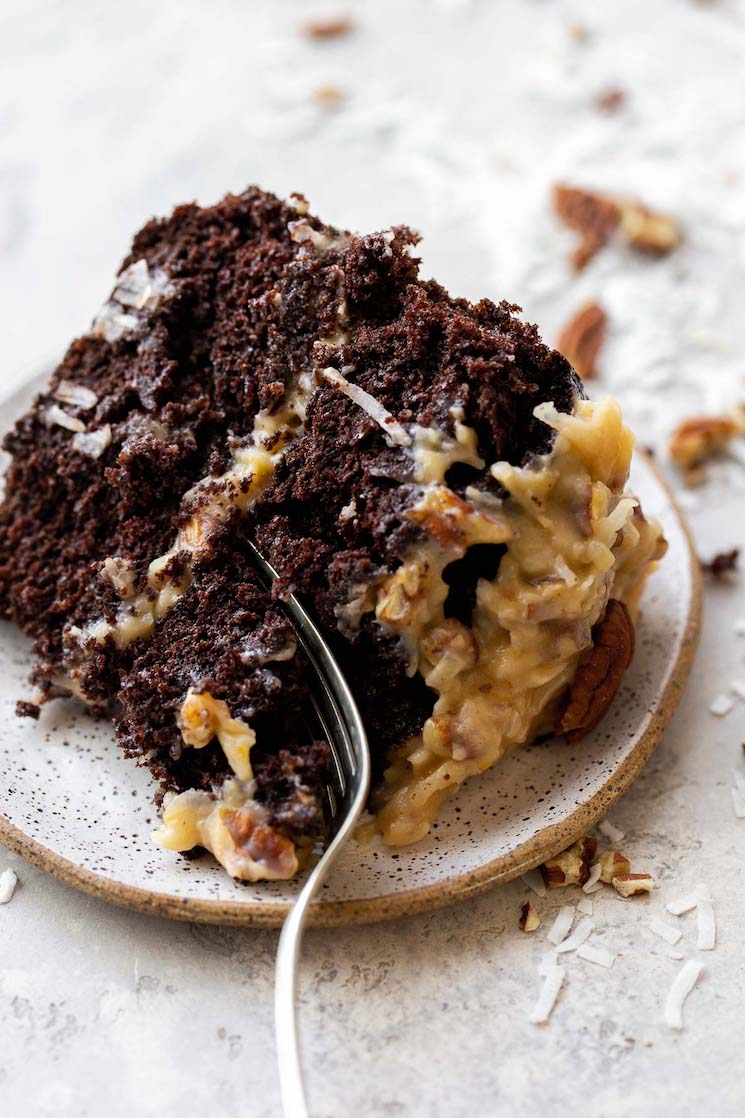 A slice of German Chocolate Cake on a rustic clay plate with a fork cutting off a piece to take a bite.