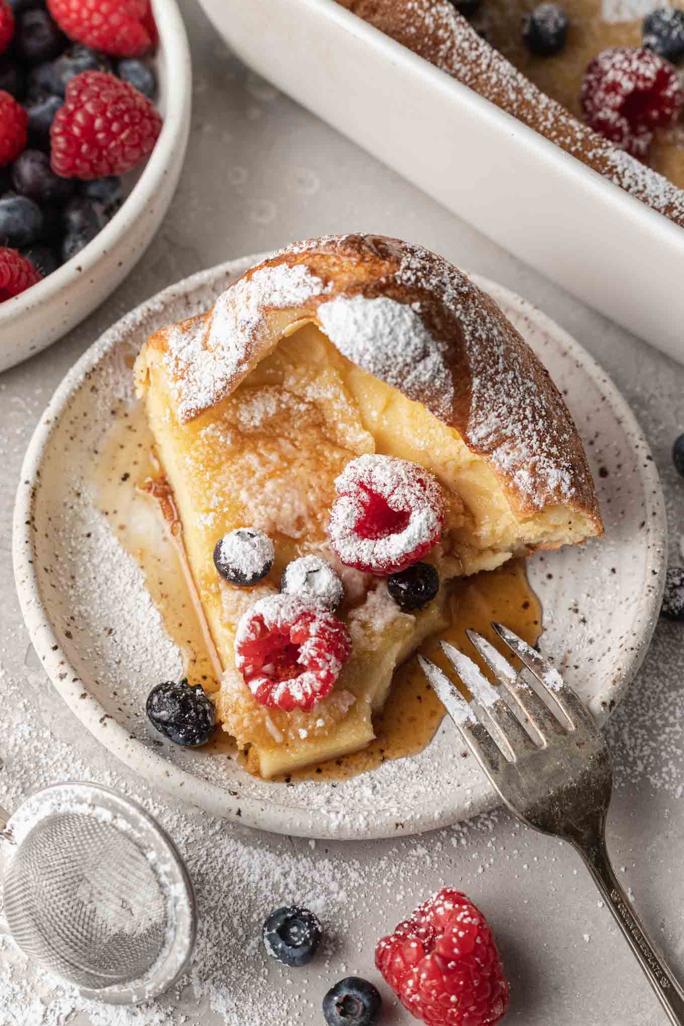 A serving of German pancakes, topped with powdered sugar, syrup, and fresh berries. A bite is missing. 