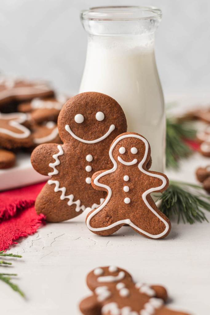 Two soft gingerbread cookies cut into the shape of gingerbread men, stood up in front of a glass jug of milk. 