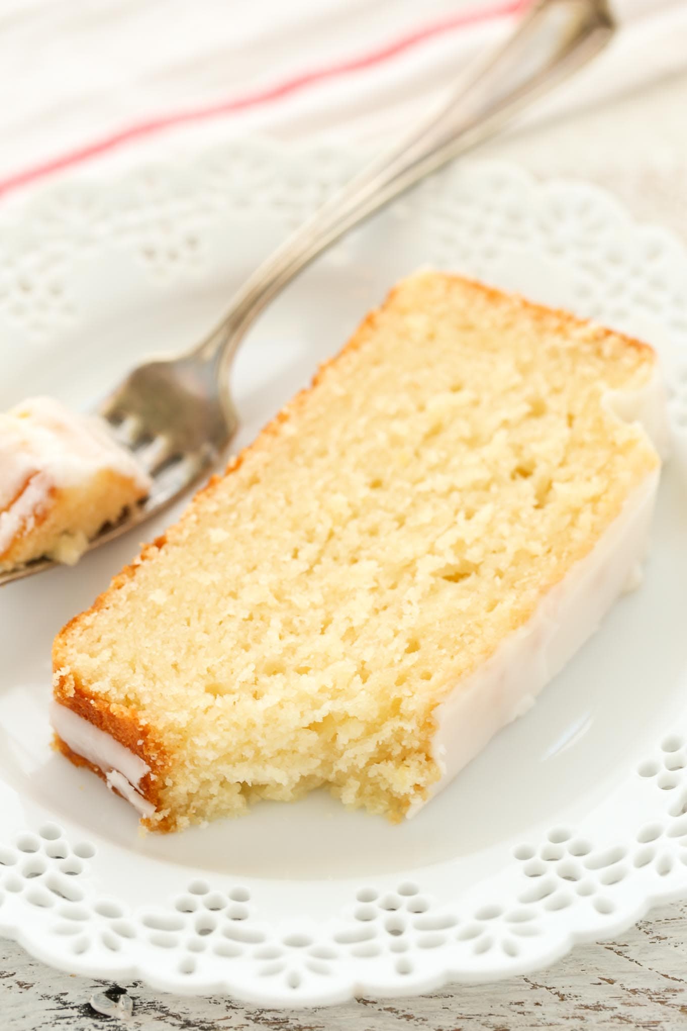 A slice of glazed lemon loaf bread on a plate with a fork. A piece has been removed and is speared on the fork. 