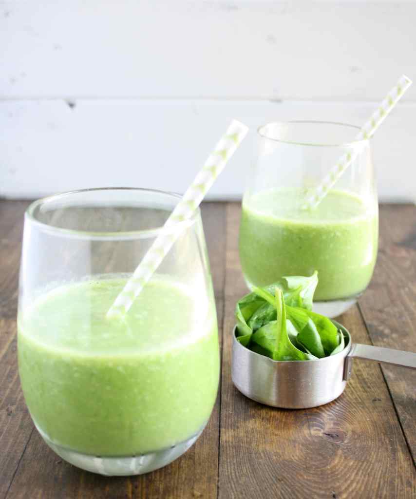 Side view of two spinach fruit smoothies with straws. A measuring cup of spinach rests between the glasses. 