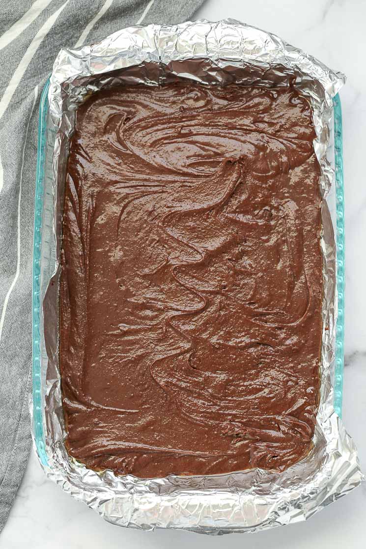 A clear pan lined with aluminum foil and brownie batter spread evenly in the pan.
