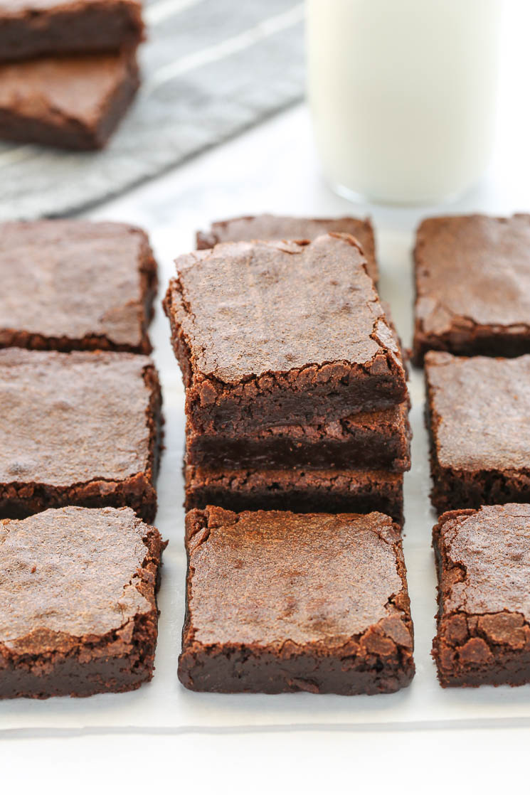 A stack of sliced brownies with other brownies around it, a glass of milk and more brownies in the background.