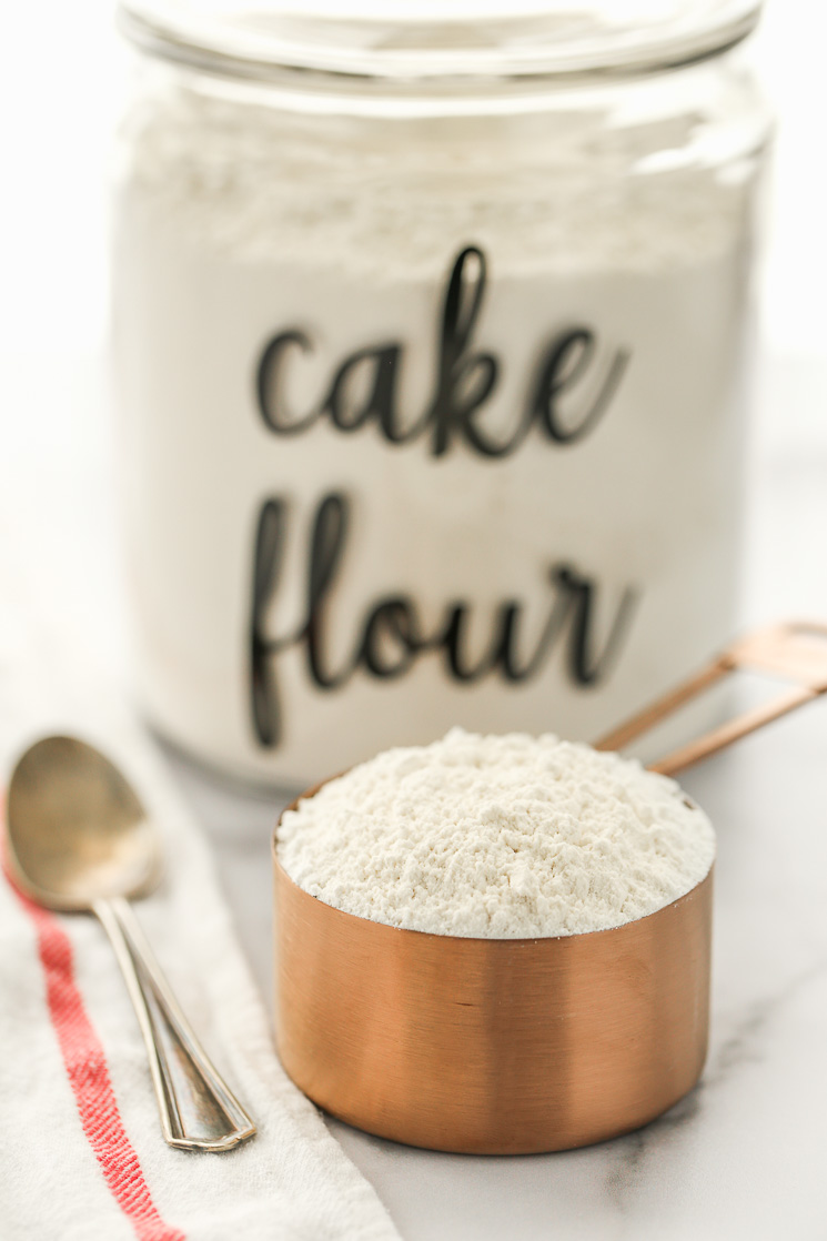 A copper measuring cup filled with cake flour and a container of flour in the background.