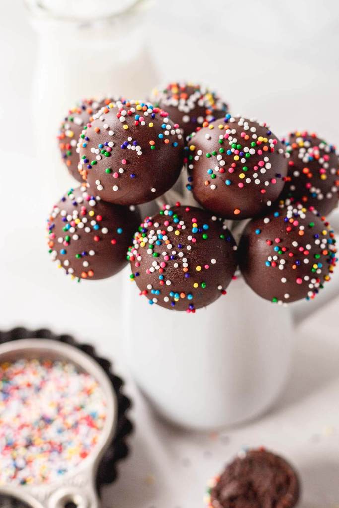 A bouquet of chocolate cake pops in a white vase. Sprinkles rest in the foreground. 